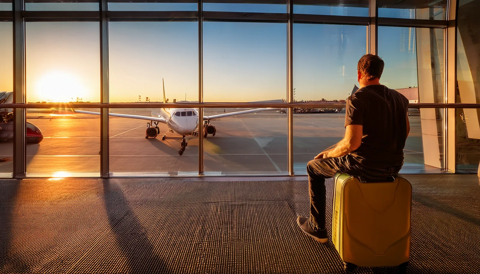 Voyageur assis sur sa valise à une porte d’embarquement, regardant un avion au coucher du soleil